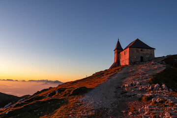 Scenic sunrise view of chapel Maria am Stein on top of mountain peak Dobratsch, Villacher Alps, Austria, Europe. Looking at Julian and Karawanks mountain range. Golden morning hour tranquil atmosphere