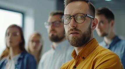 Obraz premium A man with glasses and a beard stands in front of a group of people. He is wearing a yellow shirt and has his arms crossed. The group of people are looking at him
