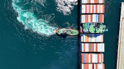 Aerial drone top down photo of tow - tug boat assisting by pulling container ship to dock to terminal port