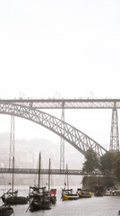 Wooden rabelos boats docked on the Douro River in Porto with wine barrels inside and the Don Luis I steel bridge with tourists strolling with umbrellas shrouded in fog on a gray rainy day.