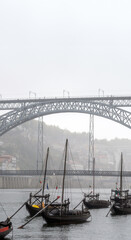 Classic wooden rabelos boats, carrying wine barrels, docked on the Douro River in Porto, beneath the Don Luis I steel bridge in the background shrouded in fog on a gray rainy day with people walking.