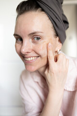 Close-up of woman in turban after washing hair looking at camera, smiling and applying cosmetic cream smear on face. Concept of morning skincare routine, anti-age care, hydration, no retouch