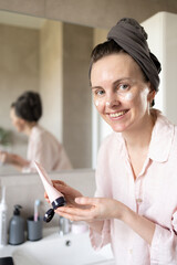 Smiling woman in turban after washing hair looking at camera and squeezing out cosmetic cream in bathroom. Concept of morning skincare routine, anti-age care. Hydration. Cream smear on face