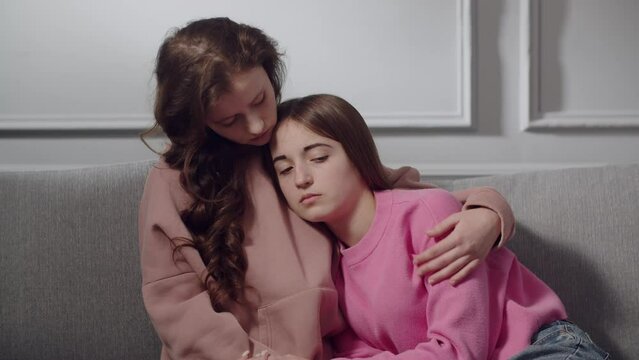 Two cute girls teenagers or students in sad emotions sit on the sofa, hug and comfort each other. A young lady looks down, leans on a friend chest, who strokes her shoulder, supports in a difficult
