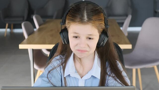 Close-up of a young girl sitting at a laptop wearing wireless headphones, chatting with her friends or answering questions during a distance learning lesson.