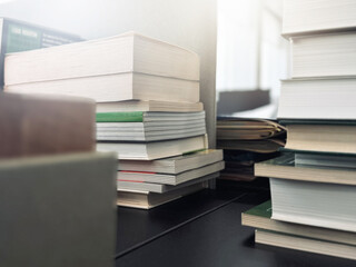 Stacks of books are on an office desk in blurred focus, illuminated by the bright daylight from the window.