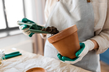 people, gardening and planting concept - close up of woman in gloves with trowel pouring soil to flower pot at home