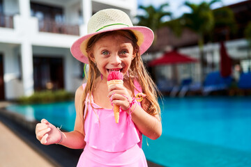 Little girl in pink swimwear and sun hat enjoys strawberry ice cream by bright blue poolside. Child with delightful expression savors summer treat, vacation ambiance, warm sunny day, leisure time.