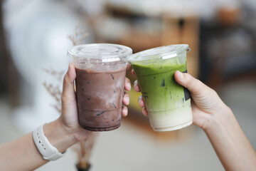 Hands of teenage girls holding glass of ice chocolate and matcha green tea together at cafe.	