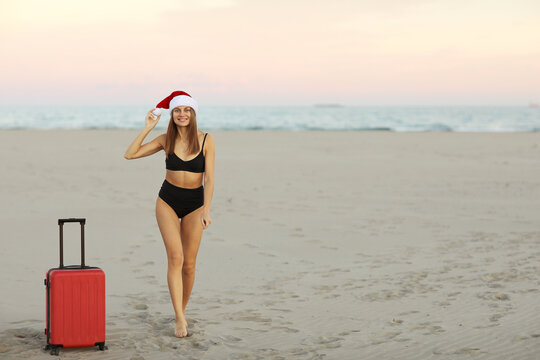 Happy young woman in red Christmas Santa Claus hat and bikini with suitcase on sand seashore shows hand for advertising booking online ticket, hot tours for travels New Year holidays.Winter vacations.
