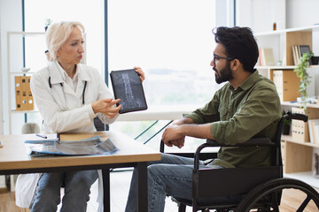 Attractive bearded young man looking on tablet with x-ray image carried by radiologist in consulting room. Confident senior specialist discussing results of diagnostic tests with wheelchair user.
