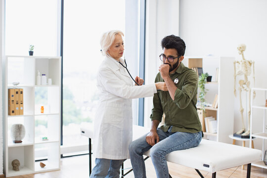 Side view of focused elderly lady in white coat using stethoscope while young man sitting on exam couch. General practitioner examining heartbeat while providing regular checkup in clinic.