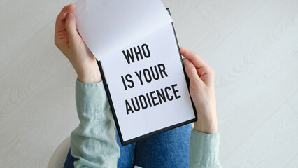 Who is Your Audience text on blank business card being held by a woman's hand with blurred background. Business concept to tell that you should know your audience.
