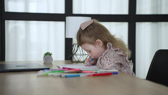 baby girl draws while sitting at table by the window at home. happy family kid concept. baby daughter learns to draw with pencils on a sheet of paper indoors. development of fine dream motor skills
