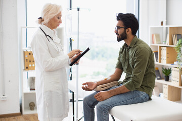 Bearded man receiving chemotherapy looking at nurse with tablet examining condition of patient. Medical specialist adjusts composition of drugs looking at electronic history of patient in exam room.