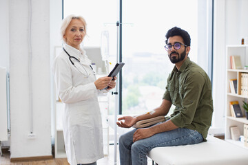 Fototapeta premium Experienced woman in lab coat using tablet for testing dropper with solution for chemotherapy for young man sitting on couch Healthcare practitioner consulting male patient with cancer in clinic.