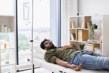 Young bearded man taking dropper with solution for chemotherapy lying on exam couch. Tired sick male in casual wear drips medicine intravenously while lying on modern light ward.