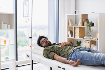 Young bearded man taking dropper with solution for chemotherapy lying on exam couch. Tired sick male in casual wear drips medicine intravenously while lying on modern light ward.