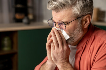 Sick middle aged grey haired man with fever and runny nose using handkerchief