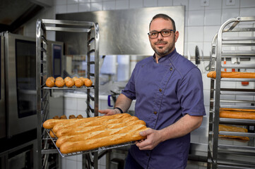 Man baker holding baguettes on tray in bakehouse kitchen