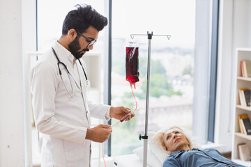 Elderly woman making blood transfusion looking at medical doctor in lab coat preparing drip. Medical specialist checking system before procedure of patient in exam room.