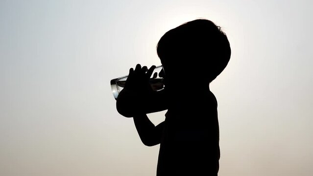 A thirsty kid drinks water from a transparent drinking glass in the hot sun during the summer midday.