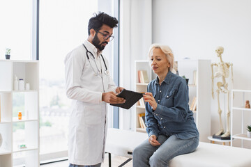 Young specialist writing instruction for taking medication correctly. Bearded male doctor and elderly female patient sitting at exam couch of modern medical center and diagnose disease.