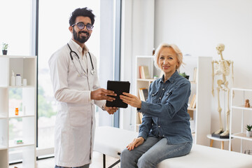 Obraz premium Portrait of bearded physician using tablet for writing electronic prescription. Senior woman describing symptoms of illness to young male doctor at modern medical office, looking, at camera.