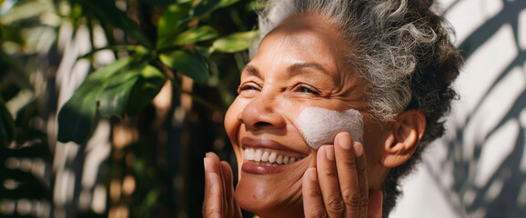 Beautiful mature black woman applying facial sunscreen lotion, standing outside in sunlight with botanical foliage