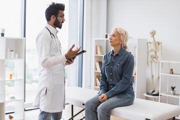 Fototapeta premium Adult bearded physician giving recommendations and using tablet for writing electronic prescription. Old woman describing symptoms of illness to young male doctor at moderm medical office.