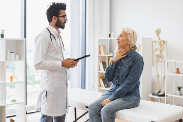 Fototapeta premium Old woman describing symptoms of illness to young male doctor at moderm medical office. Adult bearded physician giving recommendations and using tablet for writing electronic prescription.