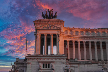 Fototapeta premium Neoclassical building at sunrise or sunset in Italy. Tall columns, pediment with sculptures, and a bronze chariot sculpture on top. Flag with green, white, and red stripes. Latin inscriptions.