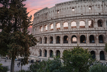 Fototapeta na wymiar Ancient amphitheater in Rome, Italy, showcasing iconic Colosseum exterior with arches, stone walls, and greenery. Tourists and locals walk near historic site at twilight, capturing serene vibe.