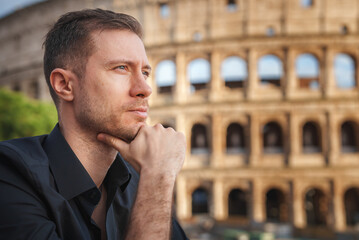 Contemplative man with light complexion and subtle stubble wearing dark shirt, with Colosseum in...
