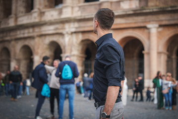 Man in dark shirt and light pants faces ancient building with arches in bustling public square,...