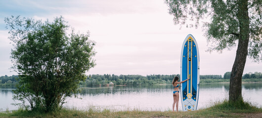 A young woman with an open swimsuit stands with a sapboard on the shore of the lake. Stand Up Paddle. SUP touring