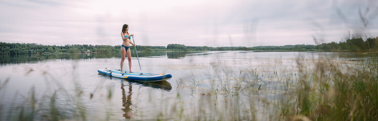 A young woman with an open swimsuit swims on a SUP board on a picturesque lake. Evening tour. A...