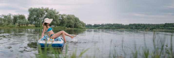 A young woman with an open swimsuit swims on a SUP board on a picturesque lake. Evening tour. A beautiful, slender girl is engaged in sap surfing on the calm water of a picturesque pond.