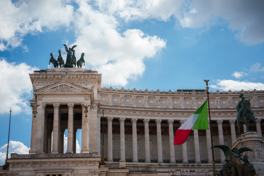 Altare della Patria, or National Monument to Victor Emmanuel II in Rome, Italy. Grand building with white columns, statues, chariot sculptures, and Italian flag. Sunny day highlights its facade.