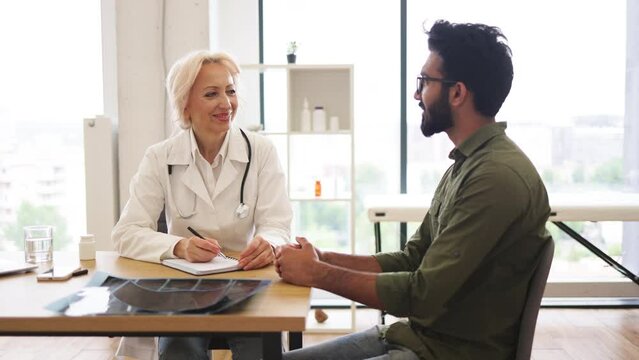 Mature elder physician giving recommendations and using notebook for writing prescription. Bearded gentleman describing symptoms of illness to senior female doctor and gesturing hands.