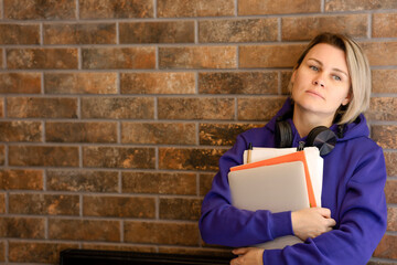 The student girl is thoughtful, looking away, leaning against a brick wall. Young blonde woman in blue hooded sweater holding laptop and notebooks, big black headphones, space for copy