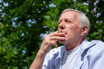 An elderly man enjoys a cigarette outdoors, with a backdrop of lush greenery, capturing a personal, reflective moment.