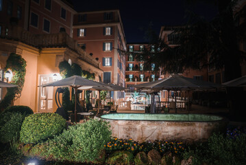 Luxury restaurant outdoor area at night with elegant garden, stone fountain, and patio covered by large umbrellas. Tranquil, exclusive setting with warm lighting and sophisticated architecture.