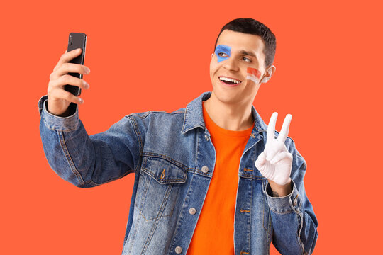 Young man with painted face and mobile phone taking selfie on orange background