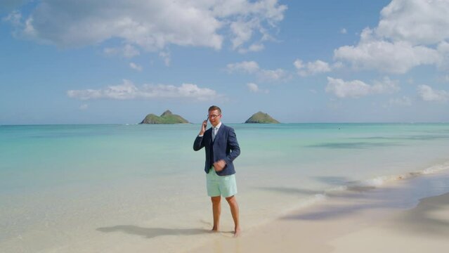 Handsome tall confident businessman making a call standing barefoot on white sand washed by iconic crystal waters of the Pacific ocean in Hawaii island. High quality 4k footage