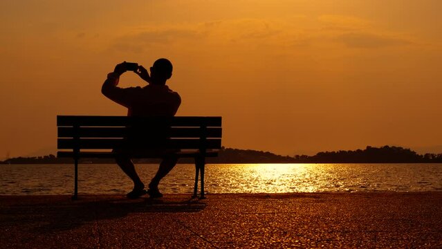 Man takes photo against evening sky. A view of man sitting on bench by the sea and takes photos of the summer sunset.