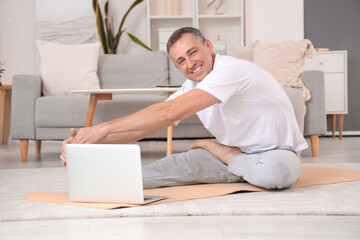 Mature man with laptop doing yoga on mat at home