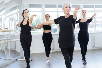Graceful elderly woman in black sportswear confidently leading ballet class with younger female dancers following her movements in spacious serene dance studio .. - obrazy, fototapety, plakaty