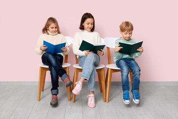 Little children reading books while sitting on chairs near pink wall