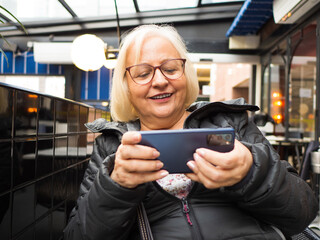 Blonde gamer grandmother surprised playing on the mobile phone on a restaurant terrace - 780151254